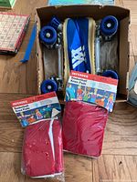 Blue roller skates with white laces and stripes paired with two boxed sets of red Defender basketball knee pads on wooden floor.