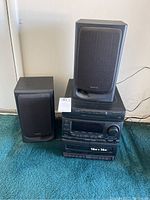 Photo showing Aiwa stereo system main unit with two speakers positioned on either side on blue carpet.
