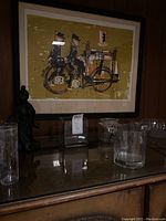 Photo showing framed wall art of two figures on a bicycle, glass decanters, ceramic figurine, and vintage bottle on a wooden cabinet with wood paneling background.