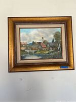 Full view of one framed landscape painting with houses near a pond and stone bridge.