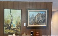 Two framed paintings displayed on a wood-paneled wall; left painting shows a tree in a landscape, right painting is an urban street scene.