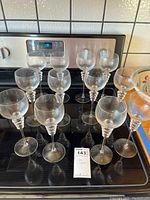 12 clear glass wine glasses with spiral stems on a black stove top.