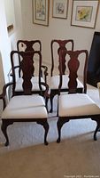 Four wooden dining chairs in dark wood with cream upholstered seats, two with armrests and two without, arranged in a room with light carpet and floral wall art.