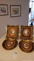 Four ornate gold-framed porcelain wall art pieces arranged with two standing and two laying flat on surface showing their decorative frames and artwork.