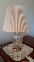 Full side view of pinwheel design glass lamp with pleated beige fabric shade on a wooden table with crocheted doily.
