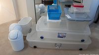 Photo showing two large clear plastic storage bins with lids and wheels, small blue basket, and several smaller clear plastic containers with lids, along with three small white plastic waste bins in front.
