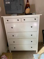 Front view of white chest of drawers with five drawers and a bottle on top.