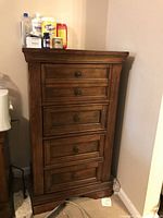 Full view of the tall chest of drawers in a corner with various items on top, showing its size and condition.