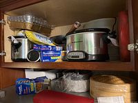 Photo of two crockpots, multiple boxes of slow cooker liners, stacked aluminum cake pans, and a red silicone baking mat stored on upper wooden shelf inside cabinet.