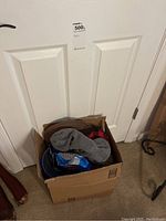 Photo showing cardboard box filled with various hats in different styles and colors, positioned against a white door.