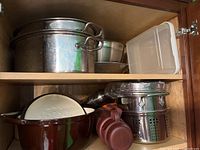 Full view of top kitchen shelves showing various stainless steel pots, bowls, colanders, white plastic container, silicone lids and a bamboo steamer
