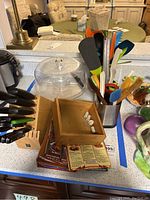 Photo showing kitchen utensils including spatulas and spoons in a metal container, knives in a wooden block, and some recipe books and pamphlets.