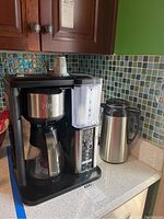 Ninja Coffee Bar machine with attached glass carafe and water reservoir, stainless steel finish with black trim, placed on kitchen counter with tiled backsplash. Thermal carafe visible beside machine.
