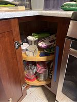 Wide-angle photo showing two-tier wooden lazy Susan inside a kitchen corner cabinet holding various storage containers, metal bowls, and kitchen textiles.