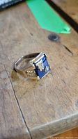 Front and angled view showing the blue rectangular stone with gold 'W' and diamond-shaped detail.