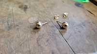 Two small spherical yellow gold ball stud earrings on a wooden surface. Each post has a butterfly clasp for securing when worn.