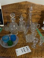 Photo showing assortment of antique glassware items including decanters with stoppers, glass trays, colored glass pieces, and small glass bowls.