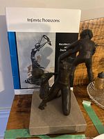Photo showing the bronze sculpture with a woman holding two children and the book "Infinite Horizons" by Jude Briggs behind it on a table.