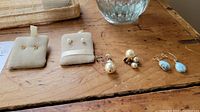 Four pairs of earrings displayed on beige earring holders and wooden surface, including two sets of pearl stud earrings, blue stone drop earrings, and clear stone stud earrings.
