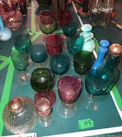Overhead shot showing all colored glass goblets, bottles, pitcher, and clear miniature lamp shade arranged on a surface with some background items.