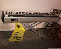 Full view of Korg SP-300 keyboard resting on adjustable metal stand with a yellow plastic bag hanging on stand frame.