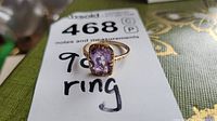 Full view of ladies 9k gold ring with rectangular purple gemstone on a white background with writing.