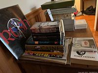 Photo showing a variety of music books stacked on a wooden furniture including a large Rush hardcover book, several other music books, a calendar, and a Red Hot Chili Peppers Greatest Hits CD jewel case.