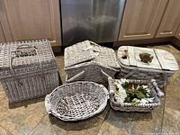 Five whitewashed wicker baskets arranged on kitchen floor: picnic basket, oval basket, two rectangular baskets including one with hinged lid, and one with fish decoration on lid