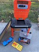 Full view of child-sized Black & Decker tool bench showing orange and black plastic construction, pegboard back with rotating rack, and two plastic tool boxes on the ground.
