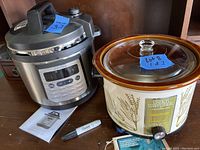 Photo showing a stainless steel electric pressure cooker with digital control panel and a 6 quart Crock-Pot slow cooker with glass lid and botanical design, including their manuals and a Sharpie marker.