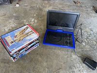 Portable blue DVD player with screen and remote, placed on a floor next to a stack of DVDs.
