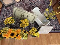 Overview showing small white wooden cradle with faux yellow flowers in metal pots, surrounded by more metal pots with bright yellow and orange faux flowers. Two decorative wooden signs placed in front.