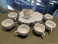 Photo showing six white opaque milk glass teacups arranged with one milk glass creamer pitcher and six biscuit plates stacked behind.