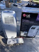 LG white portable air conditioner unit with exhaust hose attached, shown outside on concrete surface next to its original box.
