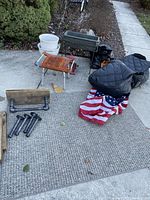 Full lot displayed outdoors on concrete, showing gray woven rug, folded American flag, industrial style shelf parts, plant pots, insulated bag, and bird feeder.
