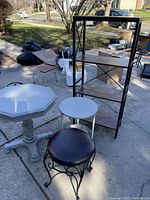 Wooden octagon table, small round metal table, black metal shelf unit, black cushioned stool outside on pavement.