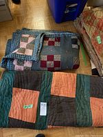 Photo showing both folded handmade quilts stacked, one with large squares in peach, green, brown, the other with smaller checkerboard patches in pink and red tones, placed on wooden floor near plastic bins.