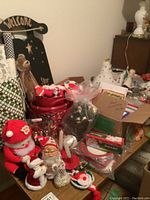 Wide view of assorted Christmas decorations, linens, boxes of Christmas cards, paper plates, figurines including Santa and angels, and small serving trays arranged on a table and nearby surfaces.