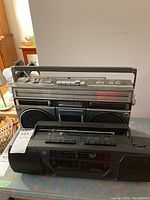Two vintage cassette players including a silver Hitachi AM/FM model TRK-7251HC with visible speaker grills and a large frequency display, and in front a black GE dual cassette AM/FM player model 3-5637A with dual cassette decks and radio tuner.