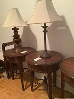 Two round Bombay style end tables side by side, each with small drawers and metal braces, each topped with a table lamp with fabric shade.