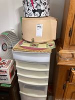 Photo showing a stacked set including a box fan, a cardboard box, placemats, a floral hatbox, and a plastic storage drawer unit.