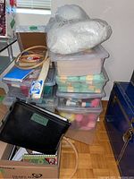 Six stacked plastic storage totes with assorted craft materials, black tote labeled 'Patterns & Crochet', wooden hoops on top, cardboard box of notebooks