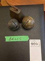 Set of three brass bells displayed on a black textured surface with a green label reading 'BELLS'. Two spherical bells and one rectangular bell with a handle are visible. A condition sticker is partially visible.