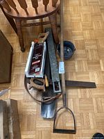 Overview of various hand tools laid on a chair including a boat hook, mitre saw in box, hand weeder, carpenter's square, shovel, and a small metal pail