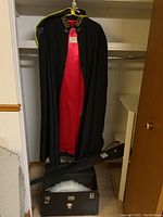 View of the black cape with red satin lining hanging in a closet, with carrying case and accessories on the floor