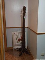 Photo of the wooden coat rack with dark finish, metal hooks with white tips, standing in a corner with paintings behind.