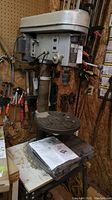 Full view of Brico drill press mounted on metal base with round adjustable table and surrounding workshop tools