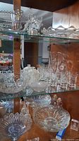 Full view of glassware and crystal items on glass shelving inside wooden cabinet, showing varied pieces including bowls, glasses, and platters.