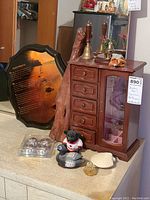 Full view of wooden jewelry box with drawers and glass door, with figurines, decorative plaque, shell, and small perfume bottle on the surface nearby.