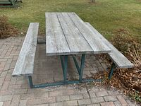 Full view of picnic table showing weathered wooden tabletop and benches attached to green metal frame on paved surface outdoors